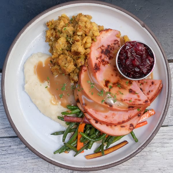 A holiday meal with sliced ham, mashed potatoes, gravy, stuffing, green beans, carrots, and cranberry sauce.