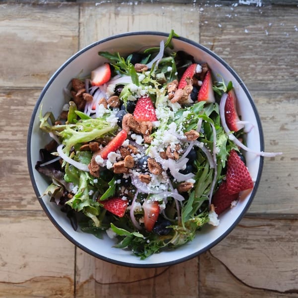A vibrant salad with mixed greens, strawberries, blueberries, feta cheese, and nuts in a white bowl.