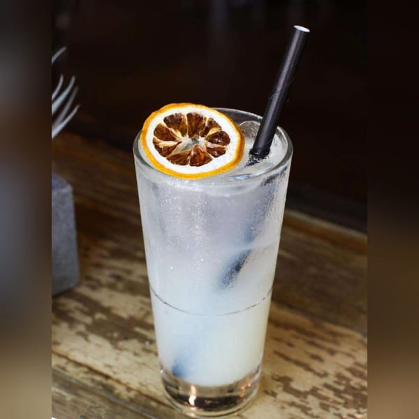 A tall, frosty glass of a clear cocktail with a dried orange slice and black straw.