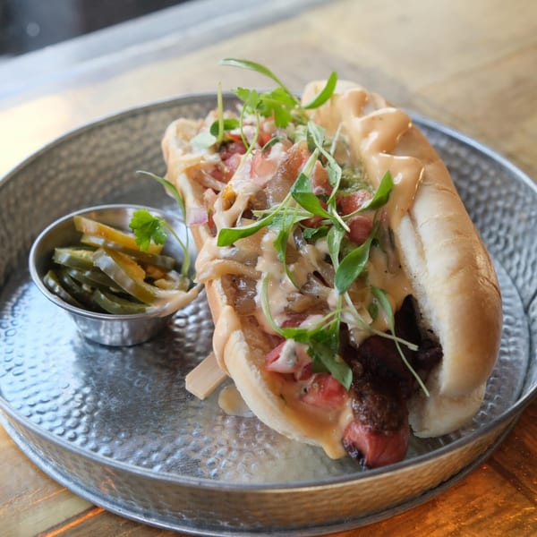 Gourmet hot dog with sauce, onions, tomatoes, and greens, served with pickled jalapeños.