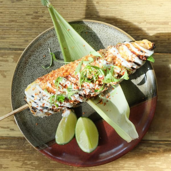 Elote, Mexican street corn, on a skewer with crema, chili powder, and microgreens, served with lime wedges.