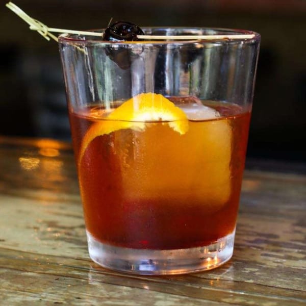 An Old Fashioned cocktail with an orange peel and cherry on a wooden bar top.