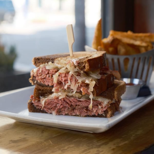 A large pastrami reuben sandwich with sauerkraut and fries on a white plate at The Corner Drafthouse.
