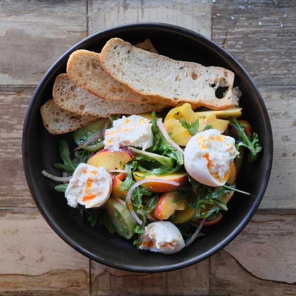 Overhead view of a fresh salad with burrata, peaches, arugula, and sliced bread in a dark bowl.