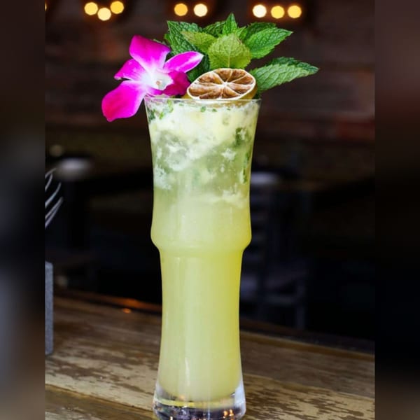 A tall glass of light green cocktail garnished with a pink orchid, mint, and dried lime.