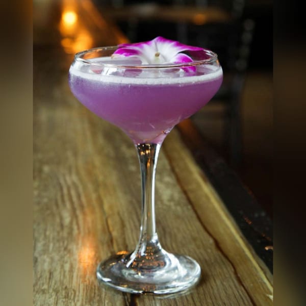 A purple cocktail with a white and purple orchid garnish sits on a wooden bar.