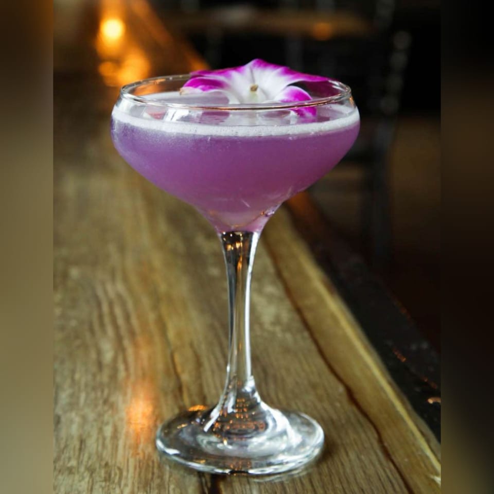 A purple cocktail with a white and purple orchid garnish sits on a wooden bar.
