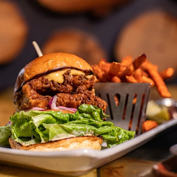 A crispy chicken sandwich with lettuce, onion, and cheese sauce, served with sweet potato fries.