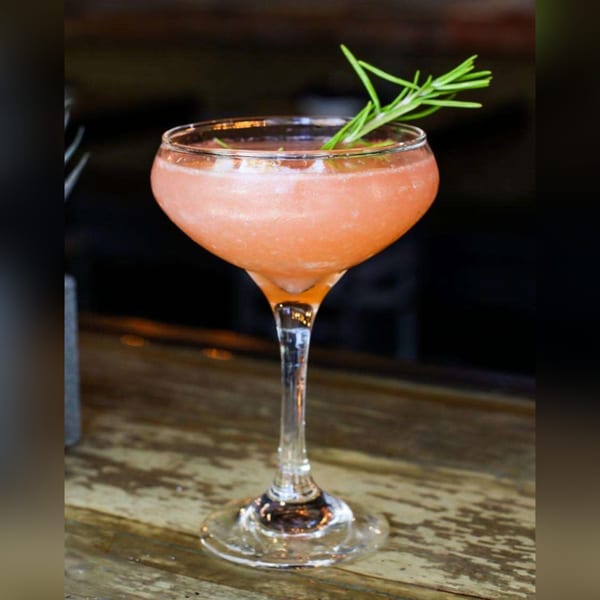 A pink cocktail in a coupe glass, garnished with a sprig of rosemary, on a wooden bar.