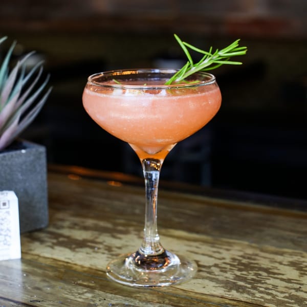 A pink cocktail with a rosemary garnish sits on a rustic wooden bar at The Corner Drafthouse.