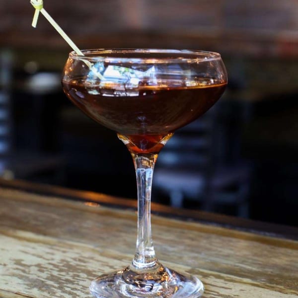 A dark cocktail in a coupe glass with a green garnish stick on a wooden bar.