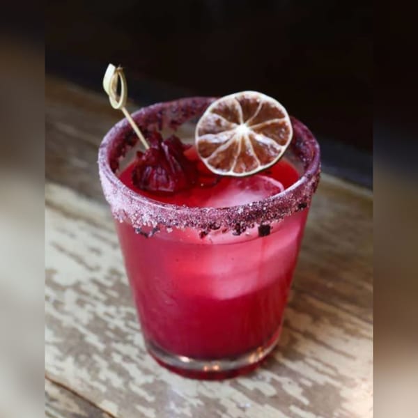 A vibrant red cocktail with a sugar-rimmed glass, garnished with a dried lime and hibiscus flower.