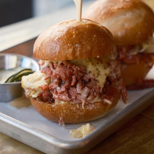 Two sliders with corned beef, melted cheese, and sauerkraut on a metal tray.