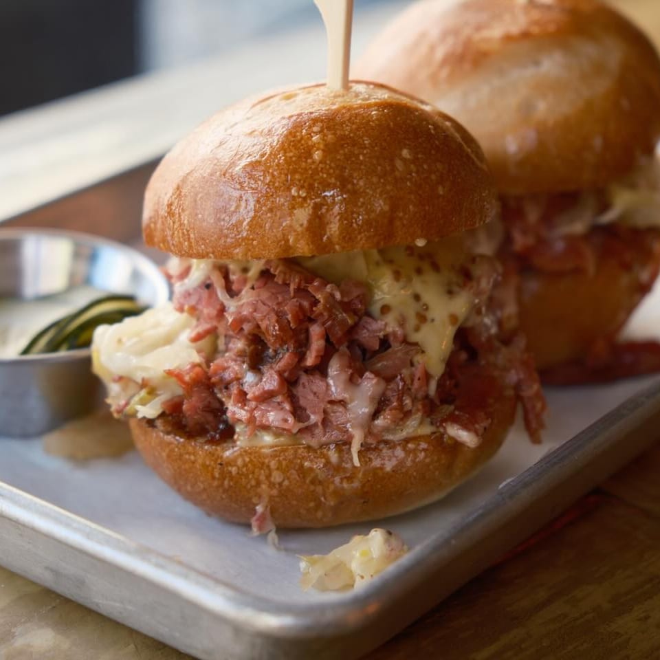 Two sliders with corned beef, melted cheese, and sauerkraut on a metal tray.