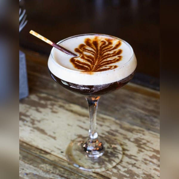 Espresso martini with a leaf design in the foam and a chocolate stick on a wooden table.