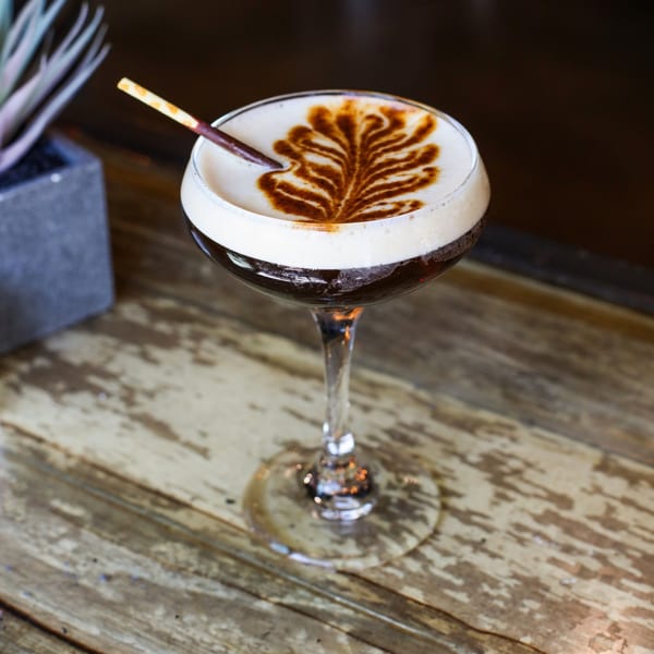 Espresso martini with foam art and a chocolate stick on a rustic wooden table at The Corner Drafthouse.