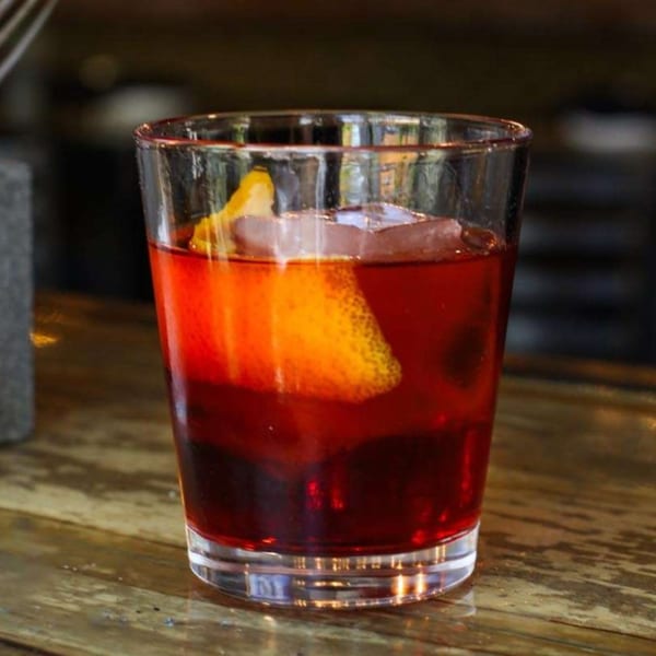 A vibrant red cocktail with ice and an orange peel garnish sits on a rustic wooden bar.