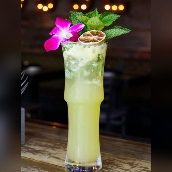 A tall, light green cocktail garnished with a pink orchid, mint leaves, and a dried lime slice.